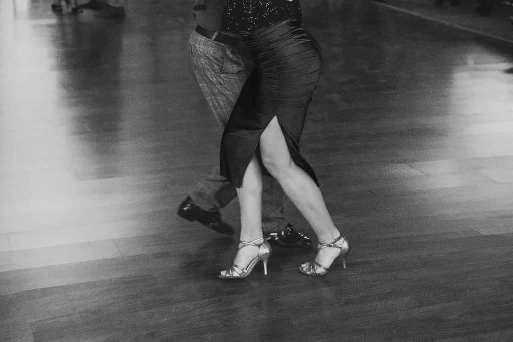 Primer plano de las piernas de una pareja bailando tango sobre un piso de madera, en una escena en blanco y negro.
