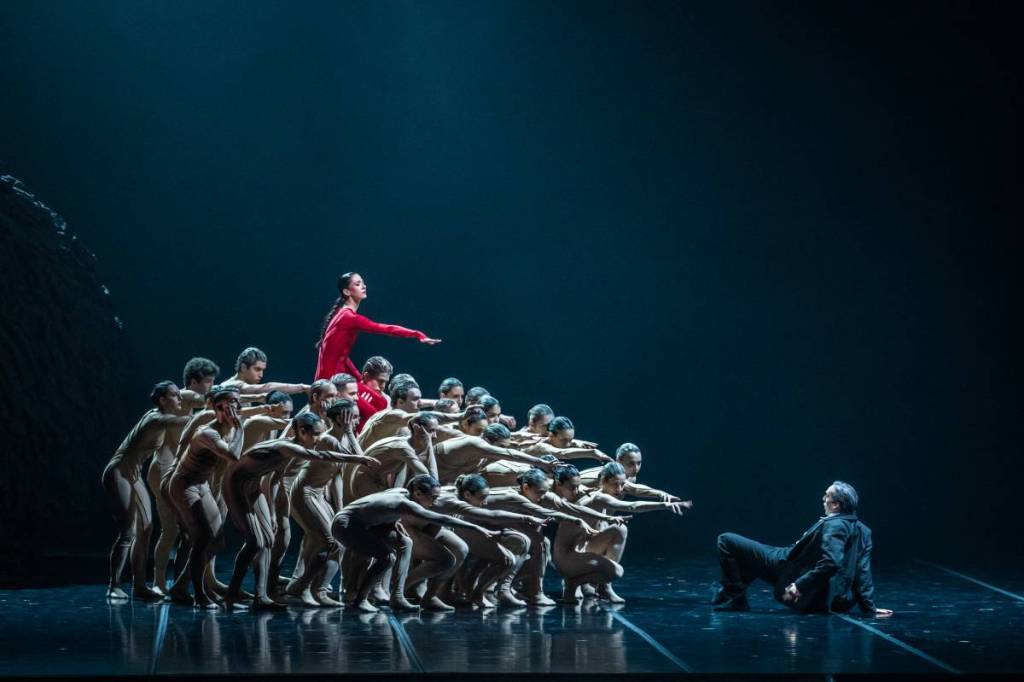 Grupo de bailarines de ballet sobre el escenario del Teatro Colón realizando una coreografía dramática con vestuario en tonos beige y rojo bajo iluminación tenue