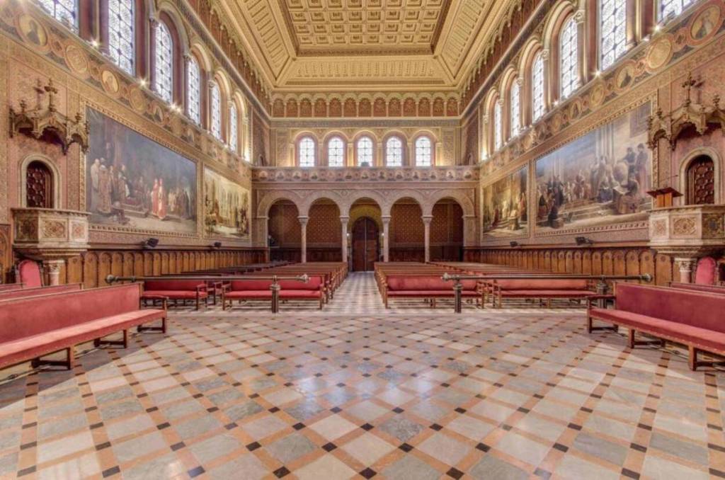 A symmetrical view of the Paraninfo at the University of Barcelona, featuring red wooden benches, historic frescoes on the walls, and a coffered ceiling.