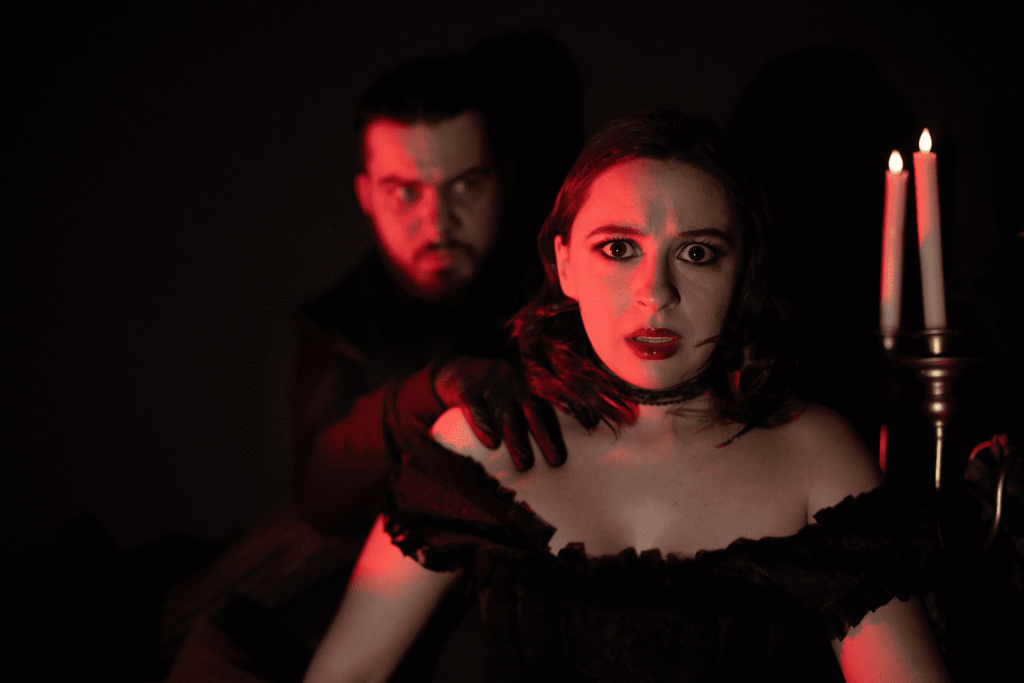Mujer con vestido negro y un hombre detrás, iluminados por una luz roja junto a dos velas encendidas.