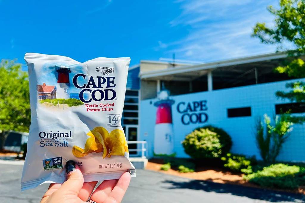 Hyannis, MA, USA - July 19, 2022: A sample-sized bag of iconic chips held up against the backdrop of the Cape Cod potato chip factory and store