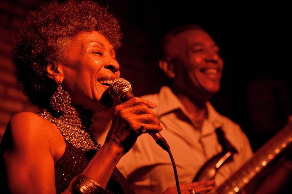 Jazz singer smiling while holding the microphone, accompanied by a guitarist under the warm and intimate atmosphere of The Jazz Room CDMX.