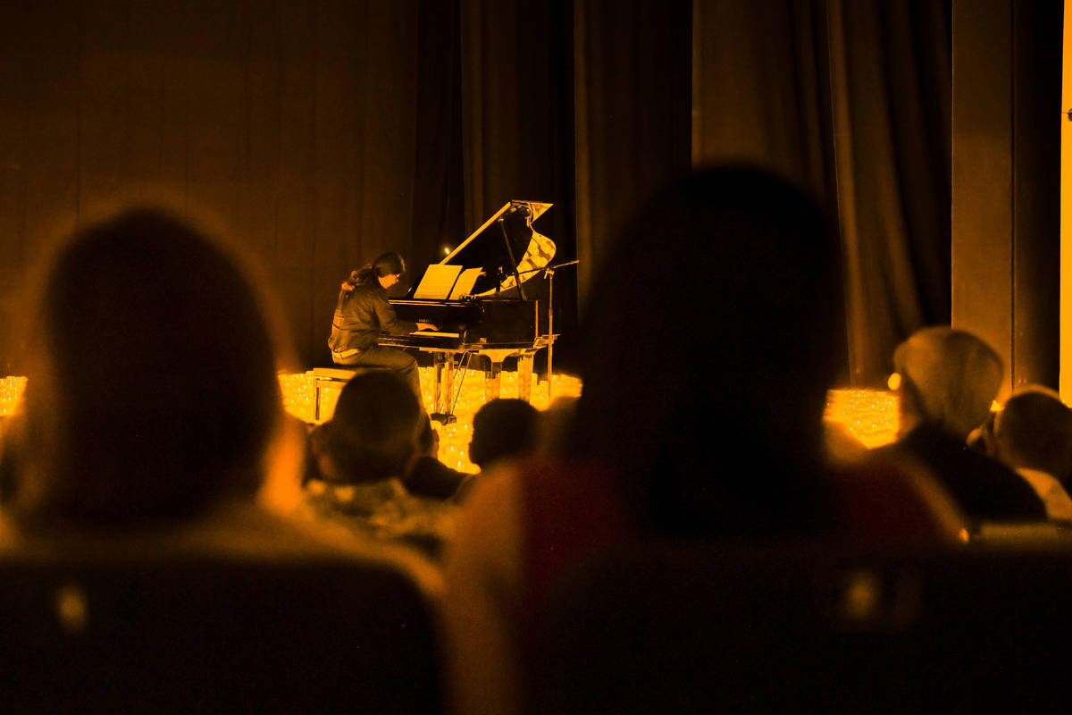 En primer plano desenfocado, varias personas del público atendiendo a un concierto Candlelight. Y, al fondo, se ve de manera muy nítica un músico tocando un piano de cola sobre un escenario cubierto de cálidas velas.