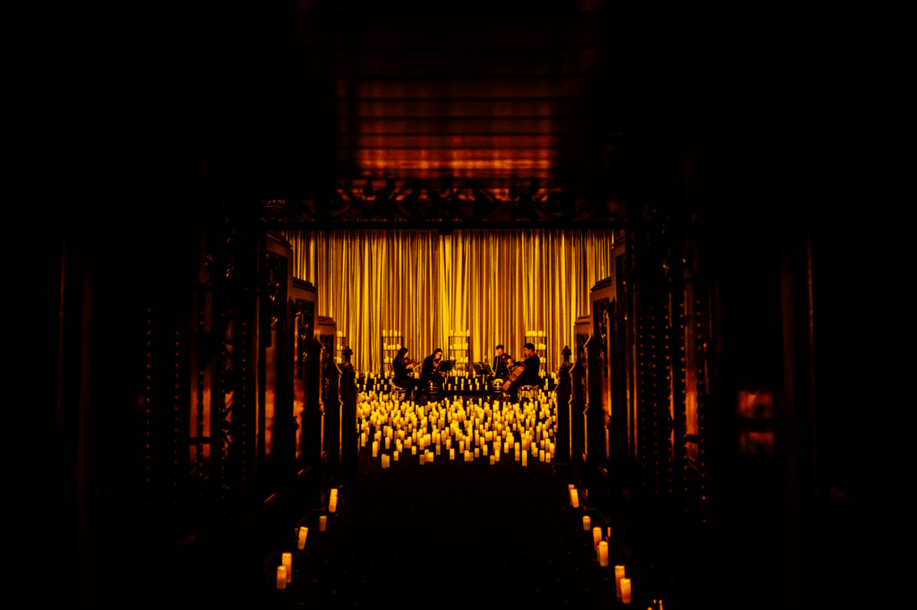 A dark aisle leading to a stage covered in hundreds of candles in the distance with a string quartet performing on the candlelit stage.