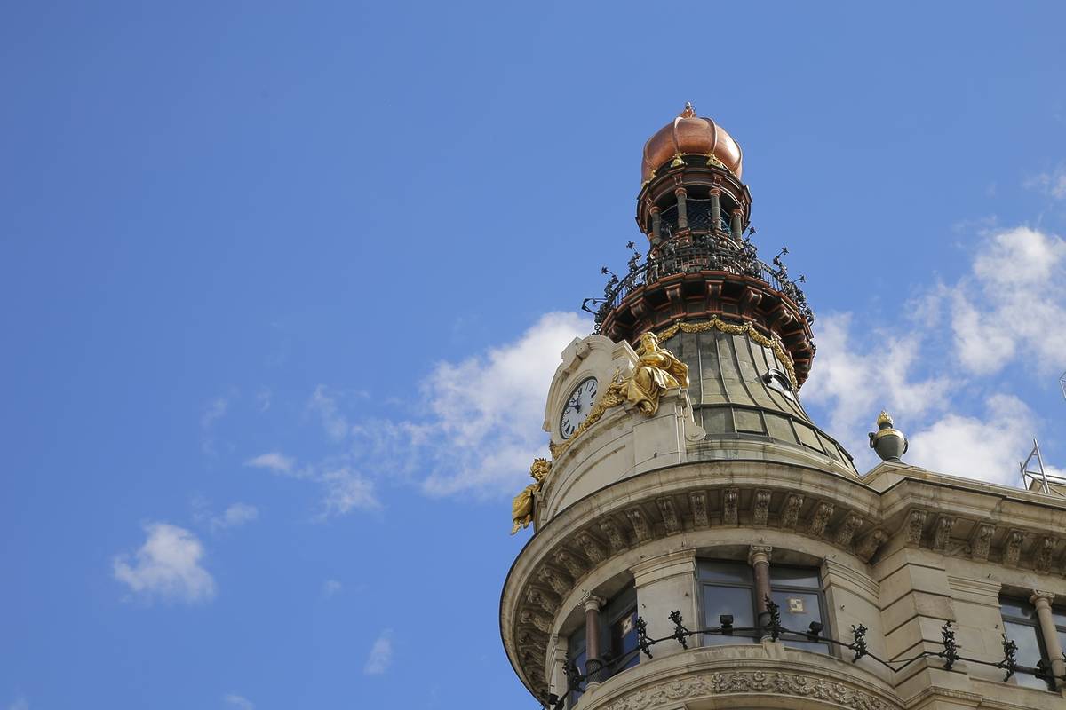 Detalle de la torre y el reloj del Four Seasons Hotel Madrid, con la cúpula de cobre y esculturas doradas sobre un cielo azul.
