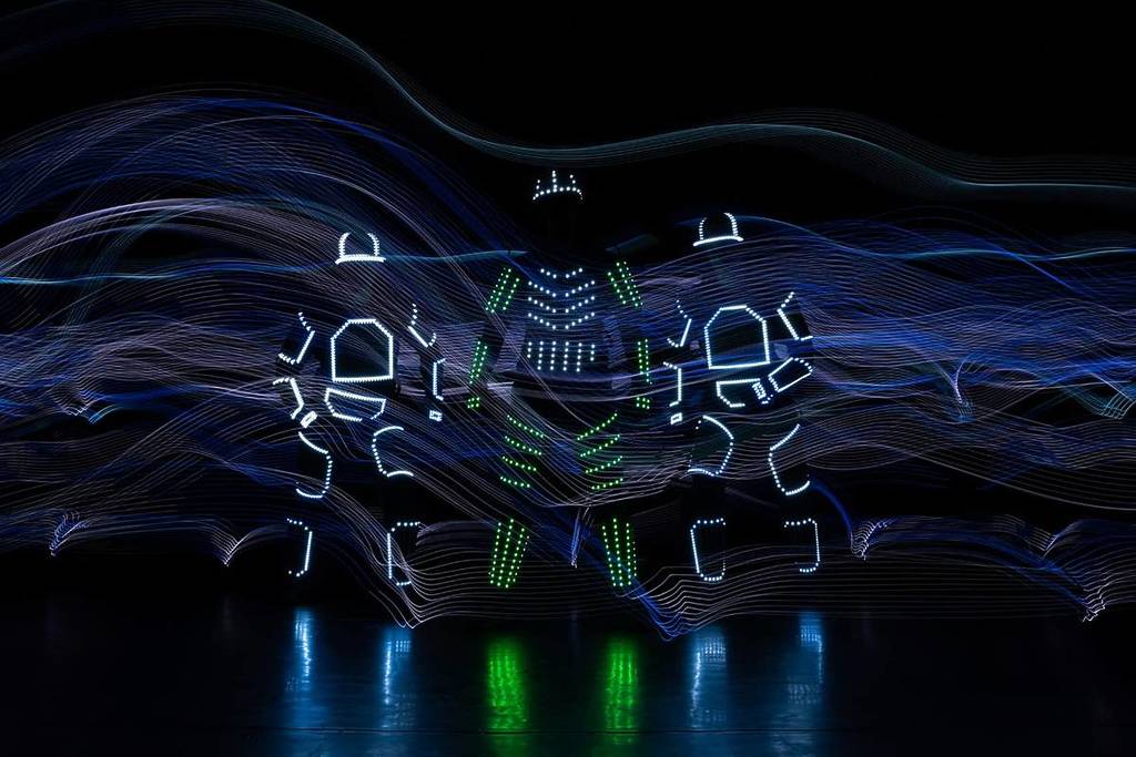 Tres bailarines con trajes cubiertos de luces LED en un escenario a oscuras.