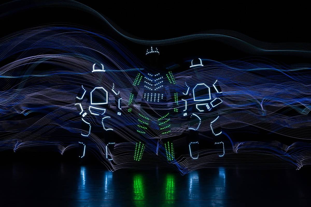 Tres bailarines con trajes cubiertos de luces LED en un escenario a oscuras.