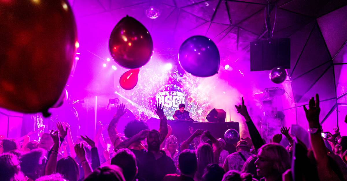 Plano general de una fiesta con globos, confeti y mucho público donde el DJ pincha sobre un escenario en Gimme Gimme Disco.