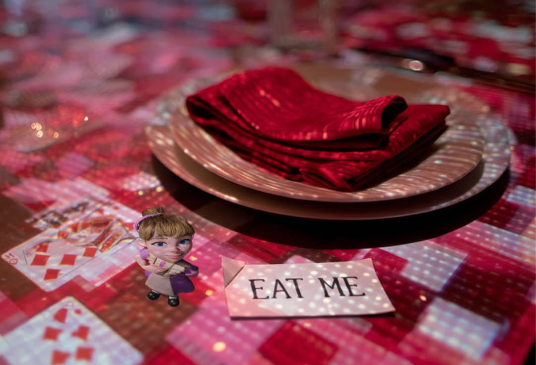 etalle de la ambientación de Sensory Feast con una mesa decorada, naipes y una tarjeta con el mensaje “Eat me”, en referencia al universo de Alicia en el País de las Maravillas.