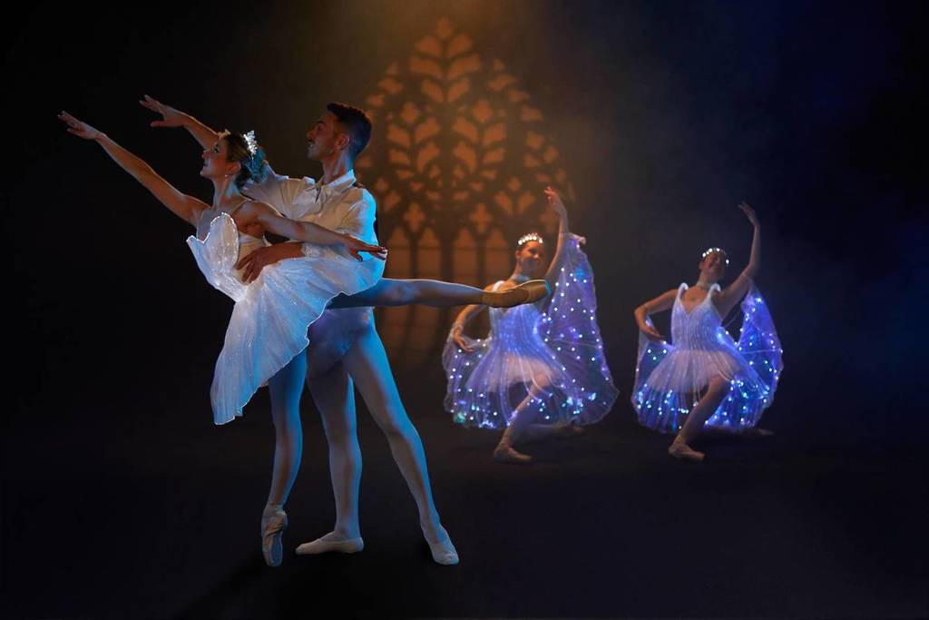 Pareja de bailarines de ballet realizando un movimiento de baile, mientras dos bailarines con trajes iluminados están por detrás