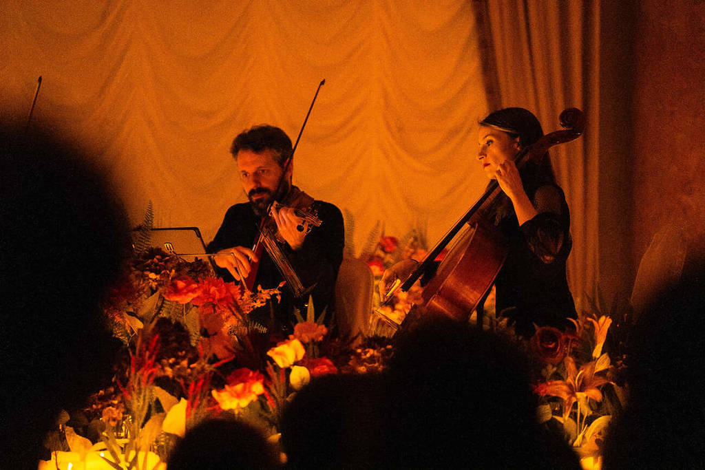 Plano medio de una violonchelista y un violinista tocando sobre u escenario cubierto de flores y velas durante un concierto Candlelight Spring.