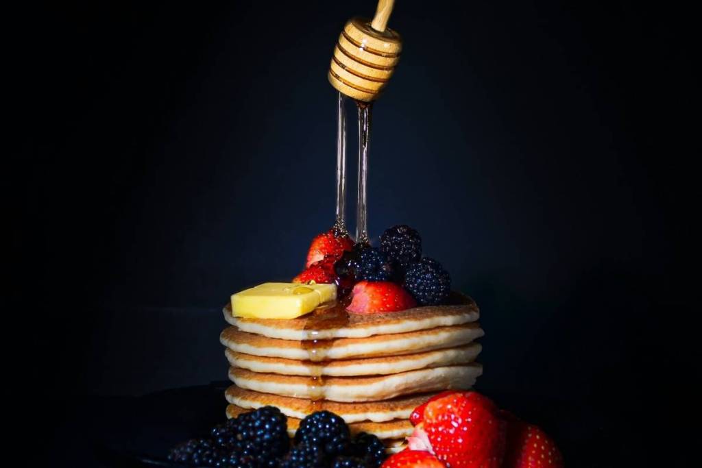 Hot pancakes stacked high with melted butter, strawberries, fresh blackberries, and maple syrup drizzled from a honey dispenser against a black background.