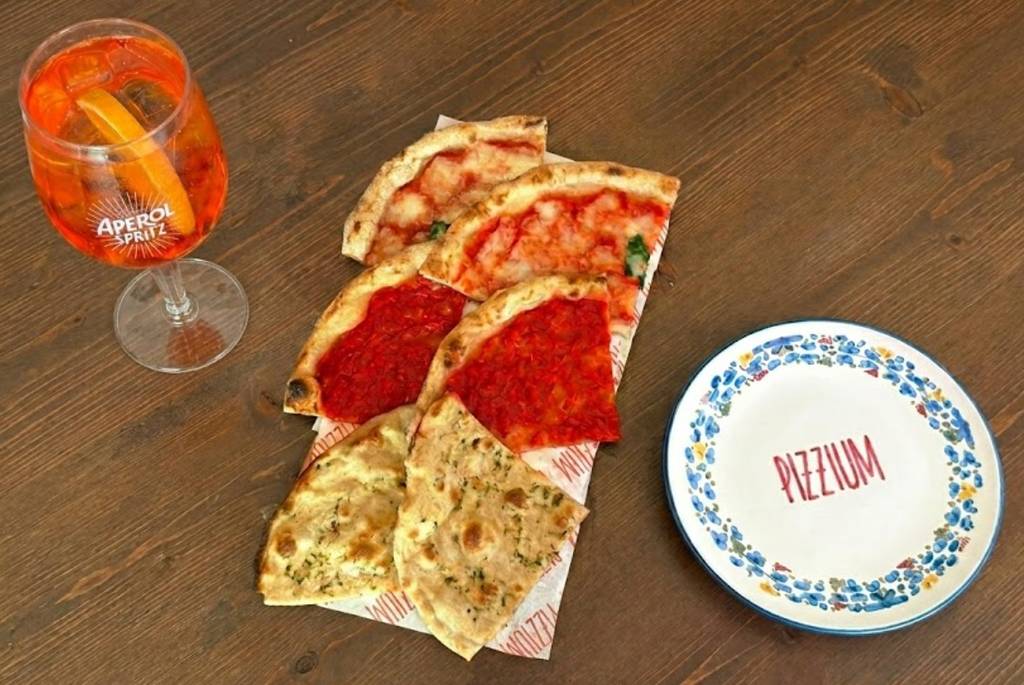 On a wooden table there is a cutting board with six slices of pizza and an Aperol Spritz, as well as a small plate with “Pizzium” written on it