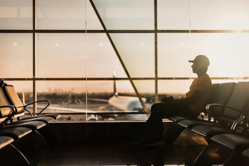 Passeggero seduto nella sala d'attesa di un aeroporto davanti alle vetrate con un aereo visibile sulla pista.