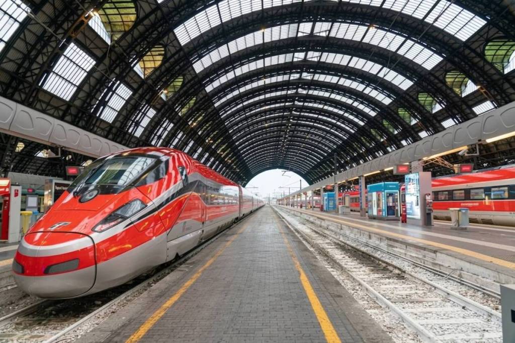 Un train à grande vitesse Frecciarossa rouge et argent à l'arrêt sur un quai de la gare de Milan Centrale, sous l'imposante structure en arcades de fer et de verre.