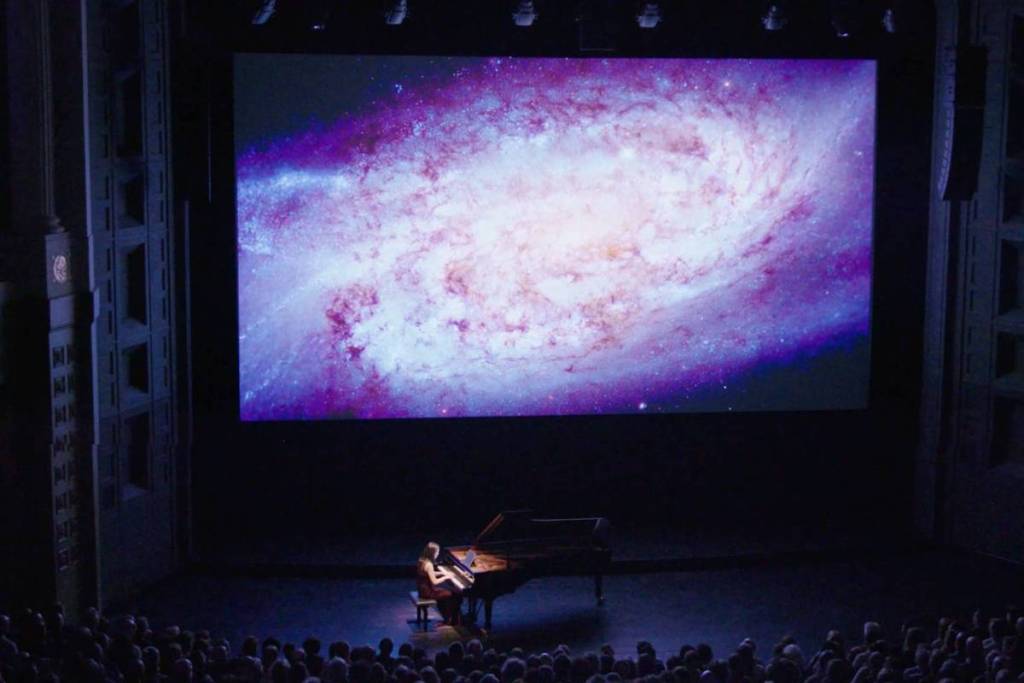 Photo of a pianist and his piano performing in front of a projection of the cosmos