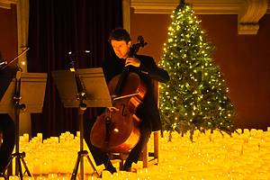 homem tocando violoncello no palco repleto de velas de LED e uma árvore de natal com pisca pisca