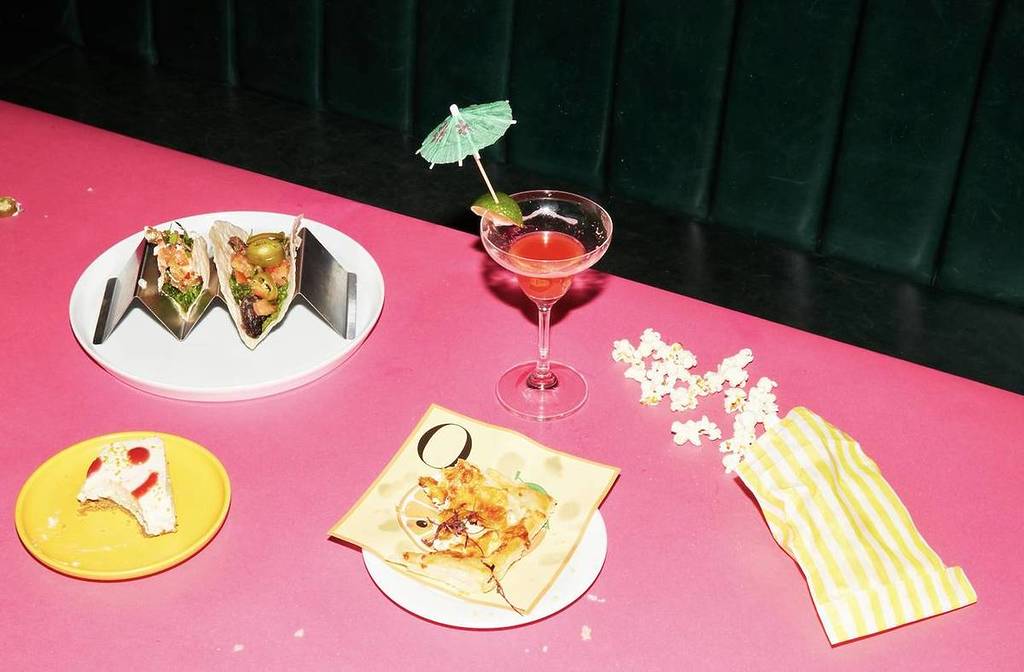 A top-down view of a pink table with cocktails and food items, shot using flash