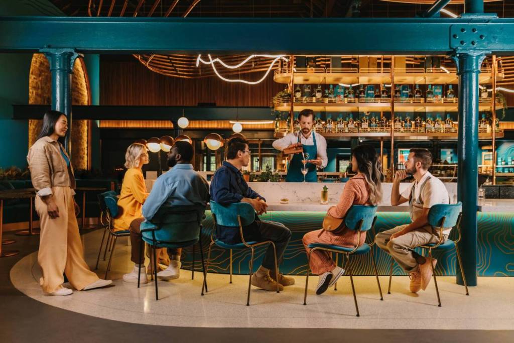 A group of people sat around a brightly lit bar, with a bartender pouring a cocktail from a shaker