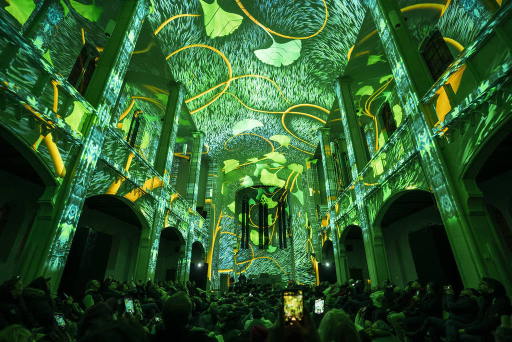 The walls and ceiling of a grand building are lit up with projections of green grass and plants. A crowd gazes up at the Eonarium Genesis show.