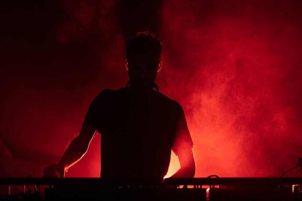 La sagoma di un DJ dietro i giradischi durante un festival underground, illuminata da misteriose luci rosse e fumo.