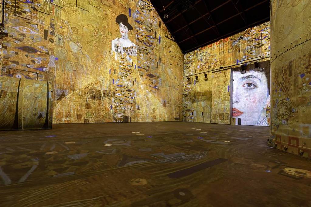 Una grandiosa proiezione artistica dorata del “Ritratto di Adele Bloch-Bauer I” di Gustav Klimt sulle pareti e le strutture di uno spazio espositivo industriale.