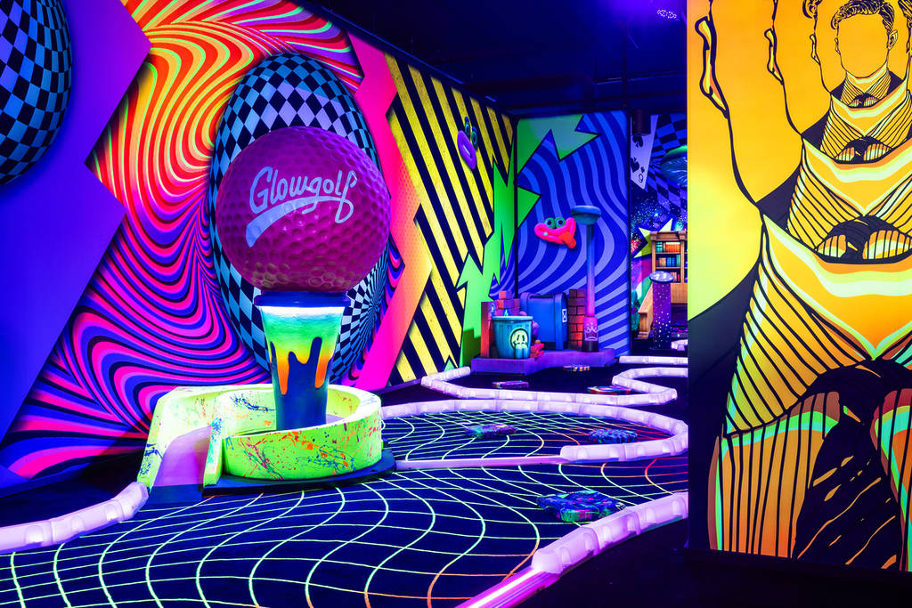 Een blacklight midgetgolfbaan met een grote roze golfbal op een tee en wanden vol kleurrijke, psychedelische neonpatronen.