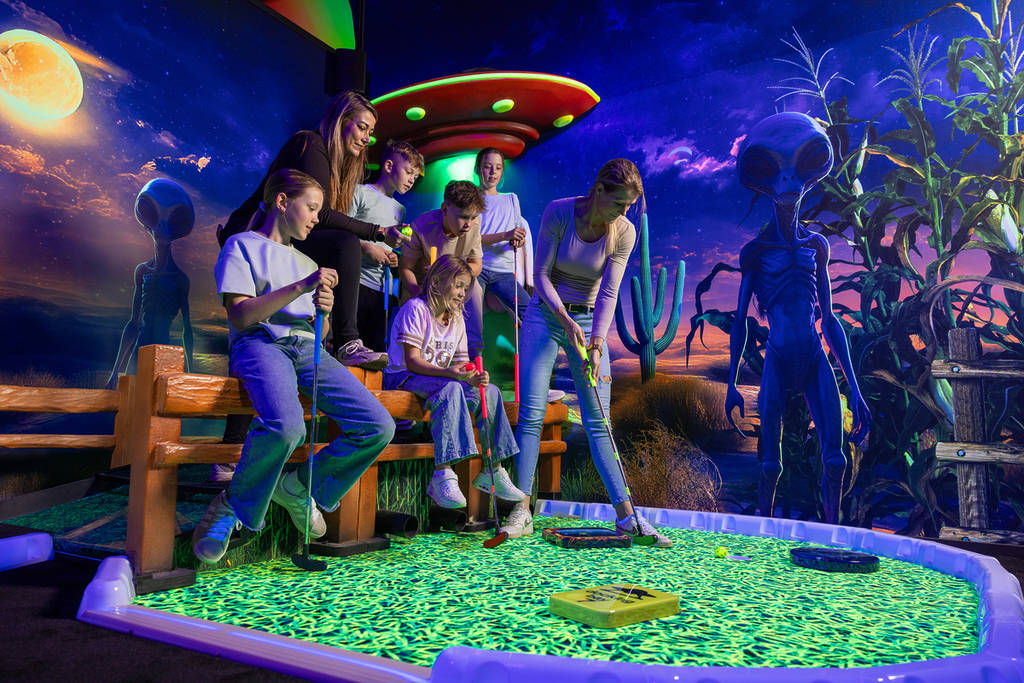 Een gezin speelt midgetgolf op een oplichtende groene baan in een ruimte met een buitenaards alien- en UFO-thema onder uv-licht.