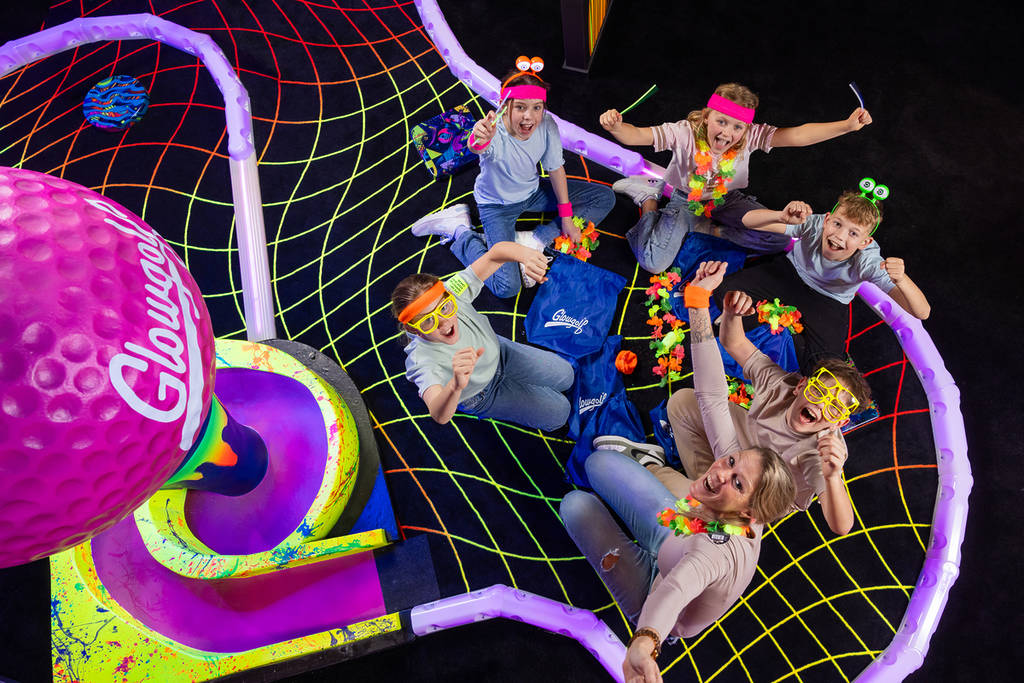 Groep kinderen en volwassenen met neon-accessoires die vanaf een vloer met spinnenwebpatroon vrolijk omhoog kijken naar de camera.