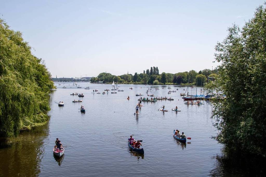 Una amplia vista desde arriba de un lago tranquilo lleno de gente en canoas, kayaks y tablas de surf de remo en un día soleado, enmarcado por frondosos árboles verdes a ambos lados.