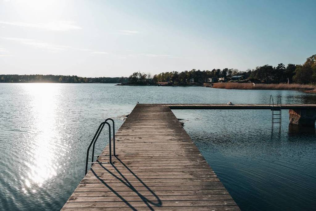 Un largo embarcadero de madera con escaleras metálicas que se adentra en un tranquilo lago recreativo con la luz del sol brillando sobre el agua y árboles en el horizonte.