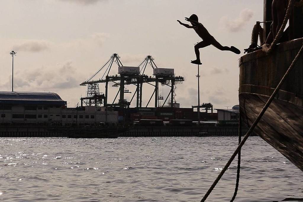 La silueta de una persona saltando al agua desde un barco de madera, con un puerto industrial lleno de grandes grúas portuarias y contenedores de carga al fondo.
