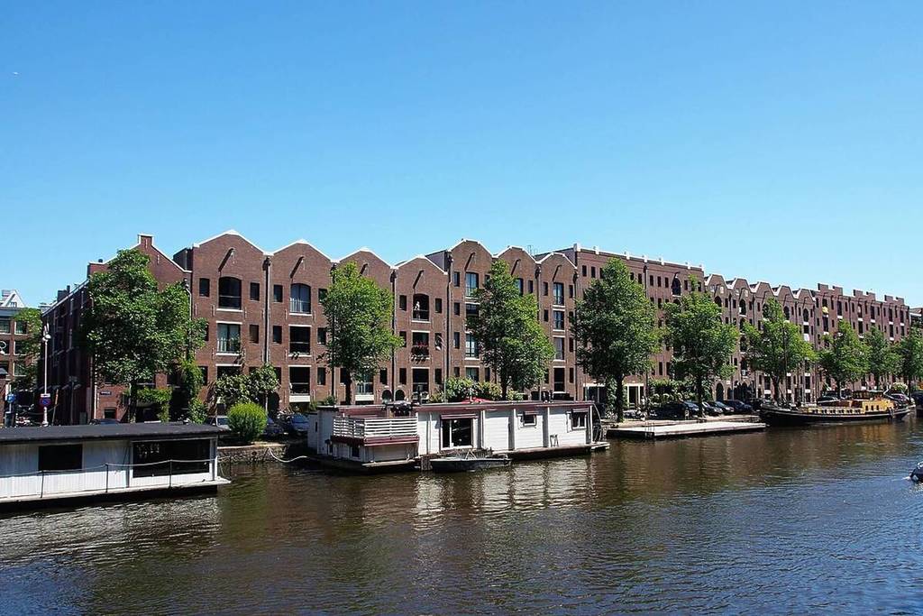 Una larga hilera de almacenes históricos de ladrillo y casas flotantes a lo largo del canal del Entrepotdok en Ámsterdam, bajo un cielo despejado y azul intenso.