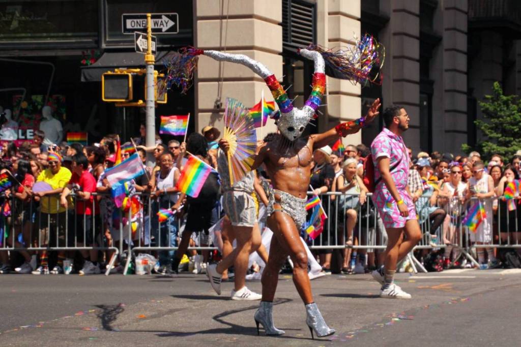 A dancing participant in a shiny silver outfit and a horned rainbow mask walks past spectators waving rainbow flags during a WorldPride parade.