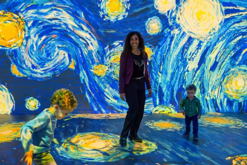 A woman and two young children experience the immersive exhibition by Fabrique des Lumières as the iconic blue and yellow brushstrokes of Vincent van Gogh’s The Starry Night are projected onto the walls and floor.