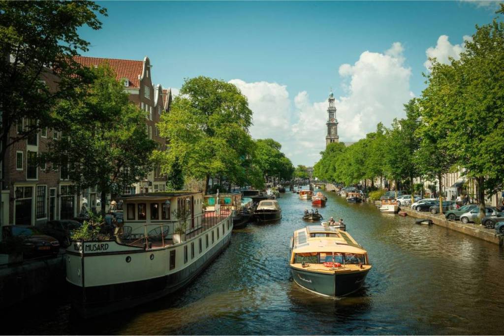 A luxury canal boat glides along the Prinsengracht with the iconic Westertoren and historic houseboats in the background on a sunny summer day.