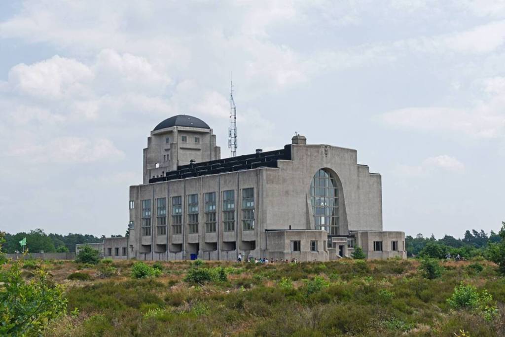 El monumental edificio principal de hormigón (Edificio A) de Radio Kootwijk, de estilo art déco, fotografiado desde la distancia sobre el brezal y los matorrales verdes que lo rodean.