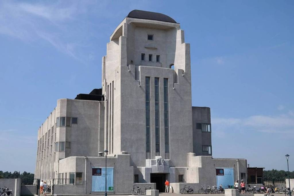 La monumental fachada y la característica torre alta del edificio art déco de hormigón de Radio Kootwijk, con visitantes y bicicletas aparcadas frente a la entrada en un día soleado.