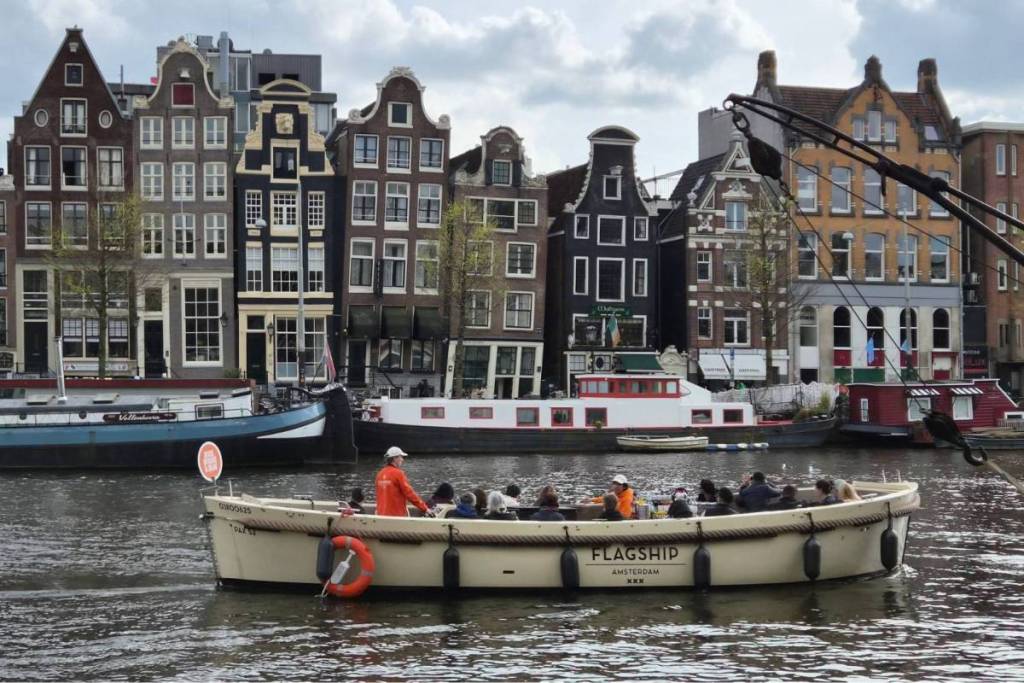 Un battello turistico aperto di Flagship Amsterdam con a bordo i passeggeri naviga su un canale davanti a una fila di edifici storici e case galleggianti di Amsterdam.