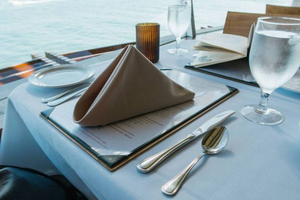 Una rappresentazione dettagliata di una tavola imbandita con un menu, un tovagliolo piegato e bicchieri d’acqua a bordo di una barca, con vista diretta sulla superficie dell’acqua circostante.