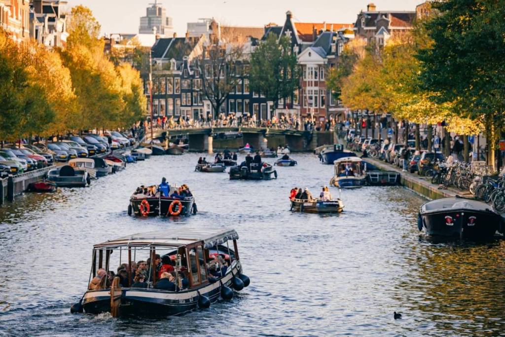 Foto orizzontale di un canale di Amsterdam con diverse barche turistiche, circondate da palazzi lungo il canale e alberi con foglie autunnali gialle.