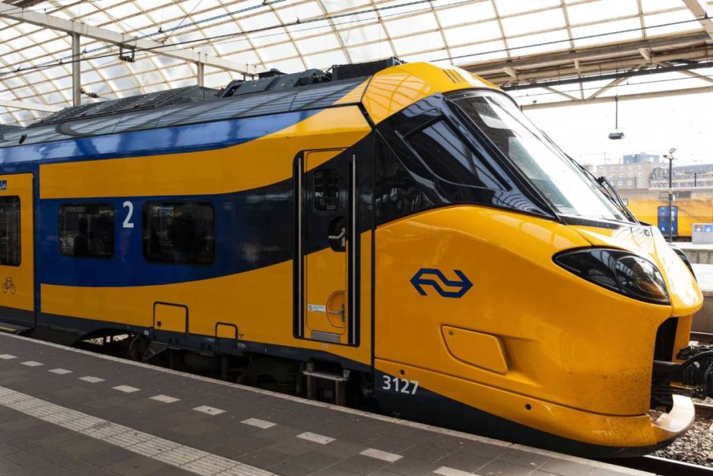 Vue de profil du nez jaune et du flanc bleu et jaune d’un train Intercity (ICNG) moderne de la NS, à l’arrêt le long du quai sous un auvent de gare en verre.