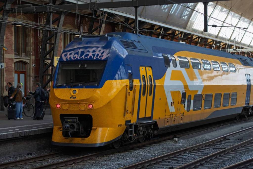 Un train à deux niveaux jaune et bleu de la NS (VIRM) est à l'arrêt le long de la voie dans une gare, avec en arrière-plan quelques voyageurs avec leurs bagages sur le quai.
