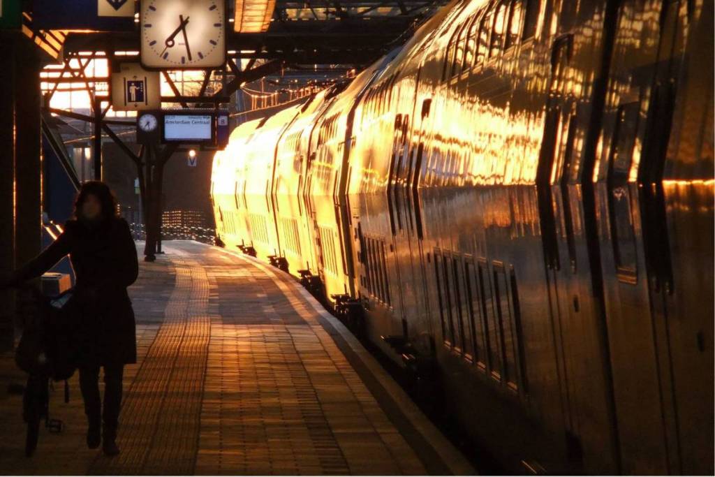Image pleine d’ambiance d’un train NS longeant le quai au lever ou au coucher du soleil, où la lumière dorée du soleil se reflète intensément sur le flanc des wagons et où la silhouette d’une personne avec un vélo se détache au premier plan.