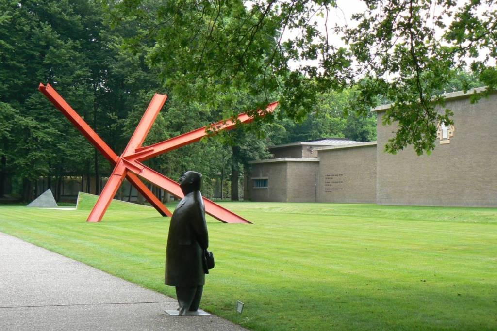 The iconic bronze statue “Meneer Jacques” by Oswald Wenckebach, overlooking a green lawn, with a large, red abstract sculpture by Mark di Suvero in the background near the museum building.