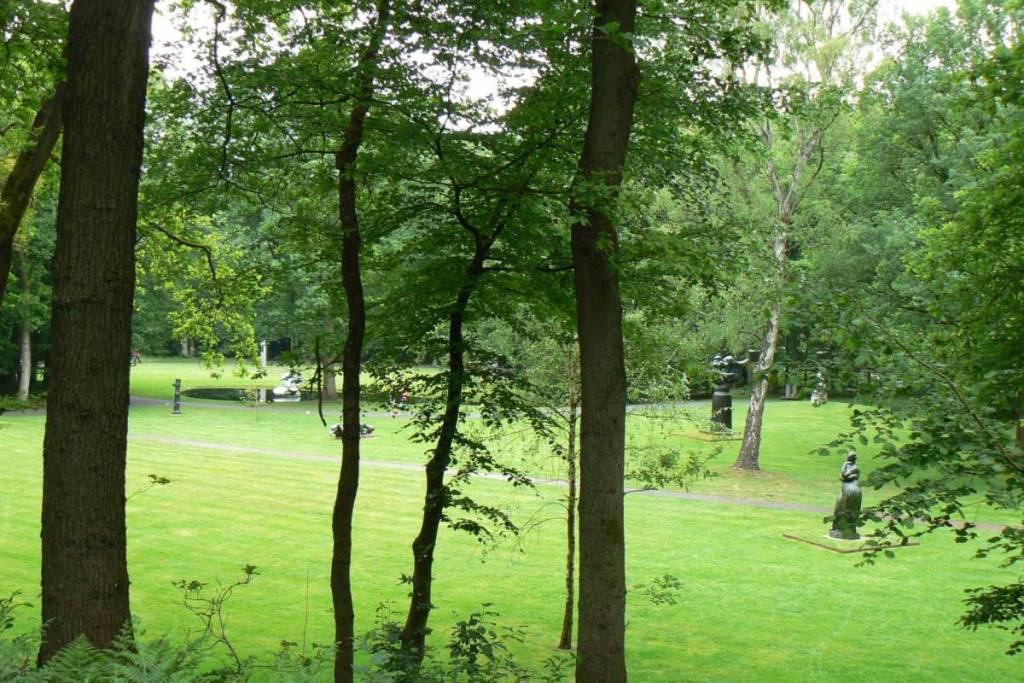 A sweeping view through the trees of a neatly mowed, green lawn in a sculpture garden, where various dark, modern sculptures are scattered across the lawns and along the walking paths.