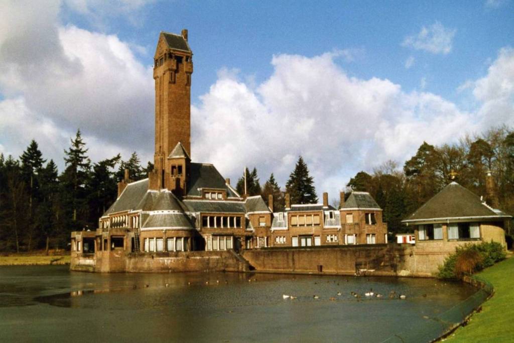 The monumental brick Sint Hubertus Hunting Lodge, with its iconic, tall tower, sits on the shore of a pond where ducks swim, beneath a blue sky dotted with white clouds.