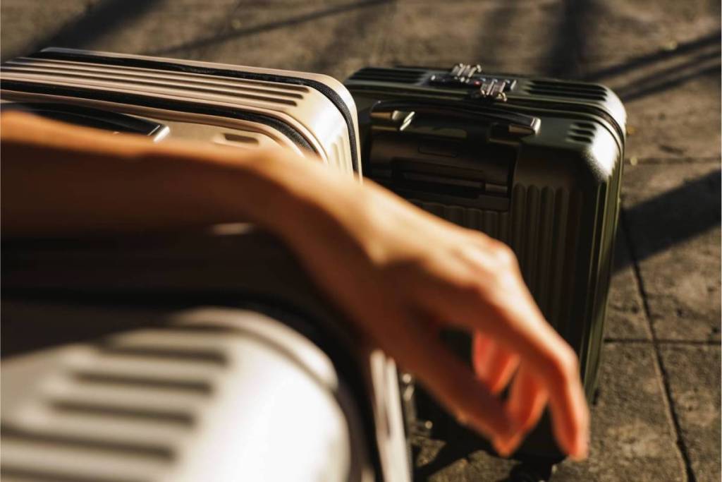 Close-up of a traveler’s hand resting on a light-colored wheeled suitcase, with a dark green suitcase next to it in the sun.