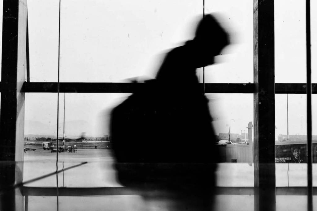 Black-and-white photo showing a blurred silhouette of a hurried traveler carrying a bag, walking past a large window in an airport terminal overlooking the tarmac.
