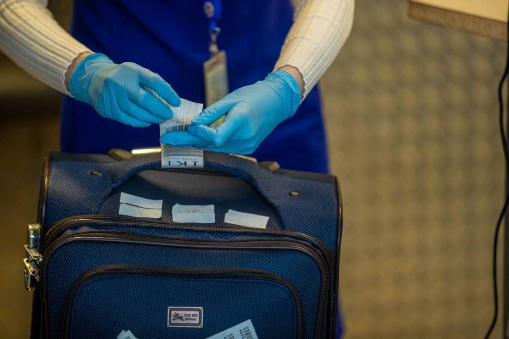 An airport employee wearing blue gloves attaches a baggage tag to the handle of a dark blue suitcase.